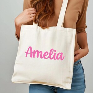 Puede incluir: Bolso tote de color crema con el nombre "Amelia" en escritura rosa brillante. El bolso tiene dos asas largas y lo sostiene una persona. El bolso est&aacute; hecho de una tela resistente, adecuada para transportar varios art&iacute;culos.