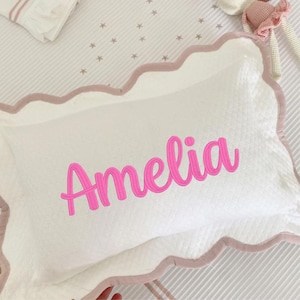 Puede incluir: Almohada rectangular blanca con un borde festoneado rosa y el nombre "Amelia" bordado en cursiva rosa brillante. La almohada tiene una textura acolchada y est&aacute; sobre una superficie a rayas.