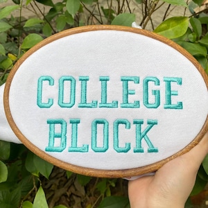 Puede incluir: Un bastidor de bordado ovalado con las palabras "COLLEGE BLOCK" bordadas en hilo turquesa sobre tela blanca. El bastidor es de madera y el fondo es follaje verde borroso.