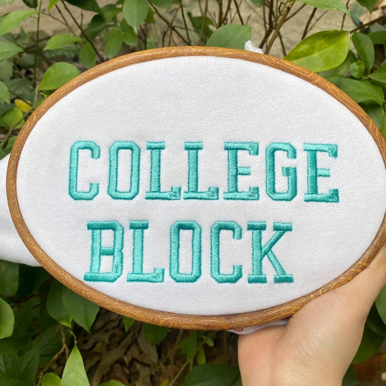 Puede incluir: Un bastidor de bordado ovalado con las palabras "COLLEGE BLOCK" bordadas en hilo turquesa sobre tela blanca. El bastidor es de madera y el fondo es follaje verde borroso.