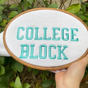 Puede incluir: Un bastidor bordado con las palabras "COLLEGE BLOCK" en hilo turquesa sobre tela blanca. El bastidor de madera es de forma ovalada y se sostiene sobre un fondo de follaje verde.