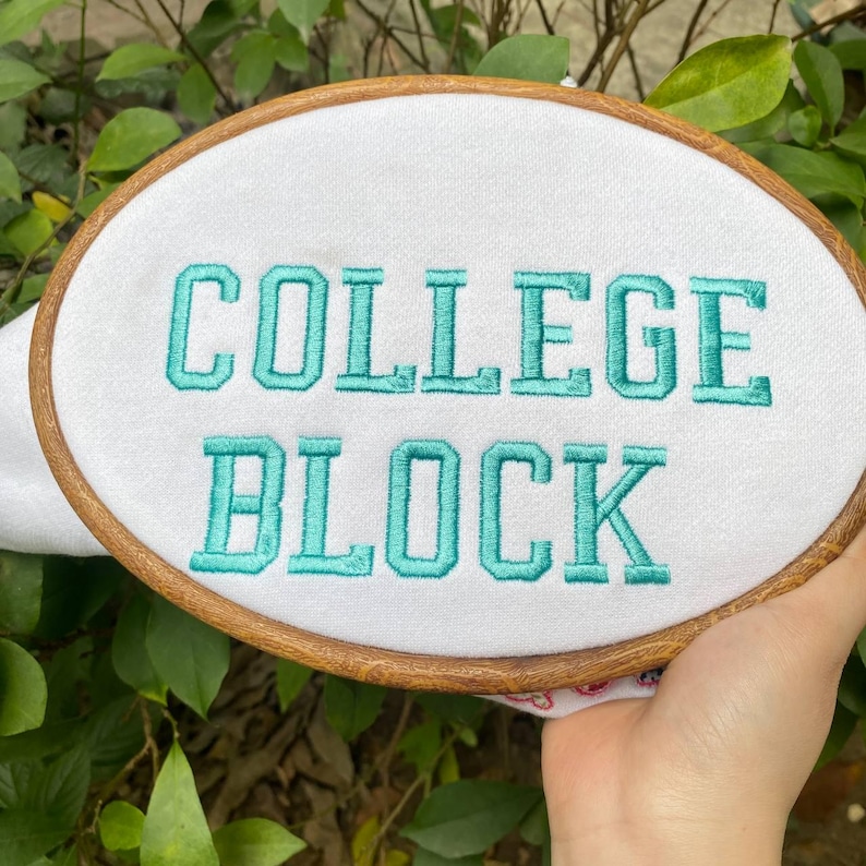 Puede incluir: Un bastidor bordado con las palabras "COLLEGE BLOCK" en hilo turquesa sobre tela blanca. El bastidor de madera es de forma ovalada y se sostiene sobre un fondo de follaje verde.