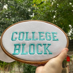 Puede incluir: Un bastidor de bordado ovalado con las palabras "COLLEGE BLOCK" bordadas en hilo turquesa sobre tela blanca. El bastidor de madera se presenta sobre un fondo verde borroso.