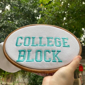 Puede incluir: Un bastidor de bordado ovalado con las palabras "COLLEGE BLOCK" bordadas en hilo turquesa sobre tela blanca. El bastidor de madera se presenta sobre un fondo de follaje verde.