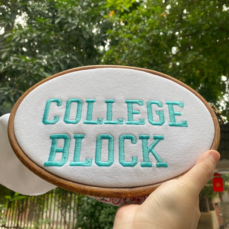 Puede incluir: Un bastidor de bordado ovalado con las palabras "COLLEGE BLOCK" bordadas en hilo turquesa sobre tela blanca. El bastidor de madera se presenta sobre un fondo de follaje verde.