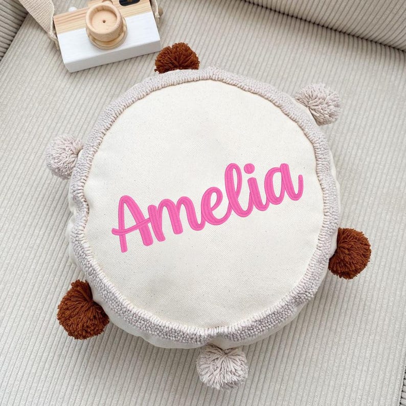 Puede incluir: Una almohada redonda de color crema con el nombre "Amelia" en rosa. La almohada tiene un borde texturizado y est&aacute; adornada con pompones marrones y beige claro. Una c&aacute;mara de juguete de madera es visible en el fondo.