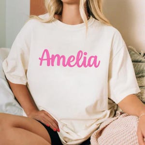 Puede incluir: Camiseta color crema con el nombre "Amelia" en escritura rosa brillante en la parte delantera. La camiseta tiene mangas cortas y un corte holgado. El texto est&aacute; centrado y destaca sobre el fondo neutro.