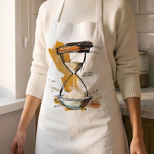 Puede incluir: Un delantal blanco con una ilustración gráfica de un reloj de arena con harina cayendo en un cuenco en la base. El diseño incluye pinceladas abstractas amarillas y marrones y el texto "SLOW DOWN MAKE BREAD" impreso en tinta negra.