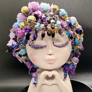 May include: A decorative ceramic face planter with closed eyes and a heart-shaped hand gesture. The planter is adorned with beaded hair in shades of purple, blue, and gold, featuring butterfly accents and glitter eyelashes. A unique and artistic home decor item.