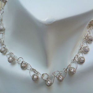 May include: A silver necklace with delicate chain links and small, intricately designed silver balls. The necklace is displayed on a white mannequin.