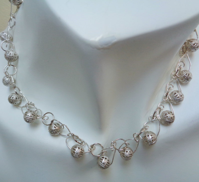 May include: A silver necklace with delicate chain links and small, intricately designed silver balls. The necklace is displayed on a white mannequin.