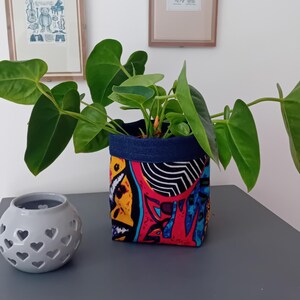 May include: A green anthurium plant sits in a fabric pot featuring a vibrant, multicoloured abstract pattern with bold red, yellow, and blue shapes, topped with a dark blue denim-like rim, positioned next to a small, round, light grey candle holder.