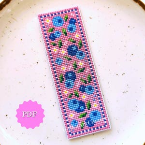 以下が含まれることがあります： ピンク色の背景に、青い花、緑の葉、小さな白い花のフローラルパターンが施されたクロスステッチのブックマーク。 ブックマークには、青と白の点線模様の白い縁があります。 テキスト「PDF」はピンク色の円にあります。