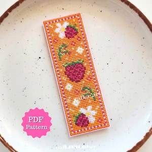 Pode incluir: Um marcador de página retangular em ponto cruz com fundo laranja, decorado com um padrão de framboesas rosa-escuras, flores brancas e folhas verdes. Um círculo rosa festonado no canto contém o texto branco PDF Pattern.