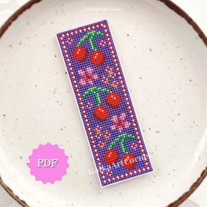 May include: A cross-stitched bookmark featuring a pattern of red cherries and pink flowers on a purple background. The bookmark has a white border with red and white accents. The text "PDF" is visible on a pink circle.