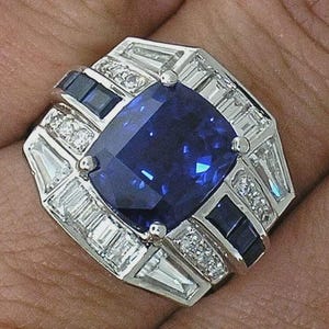 May include: A square-cut, deep blue gemstone ring with a silver setting. The ring features a large central stone, surrounded by smaller, clear and dark blue accent stones. The design is geometric and elegant, with a vintage or art deco style.