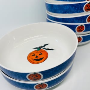 May include: A stack of white ceramic bowls with a blue mottled exterior band. Each bowl features a small orange jack-o'-lantern graphic with black bat wings on the interior base and a repeating jack-o'-lantern pattern on the outer blue rim.