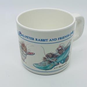 May include: White ceramic mug featuring illustrations of Peter Rabbit and friends. The mug has a blue border with the text "PETER RABBIT AND FRIENDS". The design includes a rabbit holding a plate and mice in a blue shoe.
