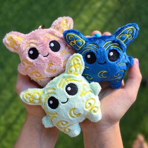 May include: Three plush toys in pink, blue, and mint green, each with embroidered yellow crescent moon and swirl designs. The toys have large black eyes and small smiles, with a cute, cartoonish appearance.