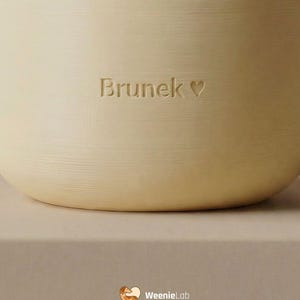 May include: A light beige pet bowl with the name "Brunek" and a heart symbol engraved on the side. A paw print is at the top. The WeenieLab logo is at the bottom.