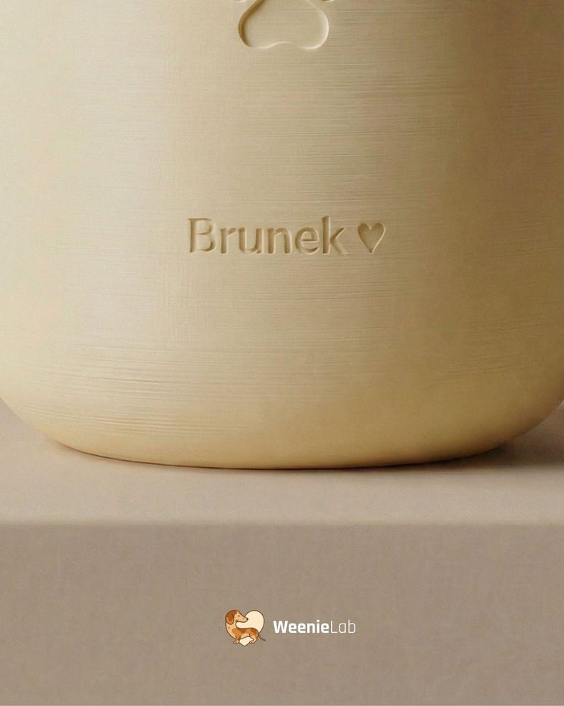 May include: A light beige pet bowl with the name "Brunek" and a heart symbol engraved on the side. A paw print is at the top. The WeenieLab logo is at the bottom.