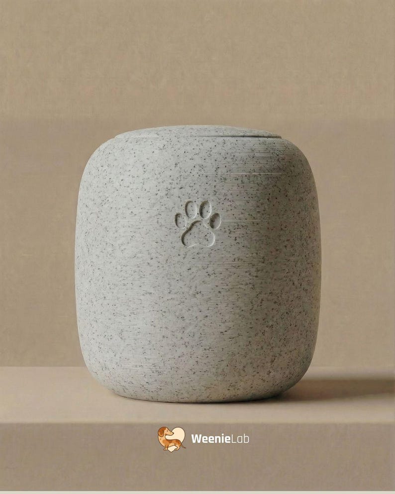 May include: A speckled gray pet urn with a paw print design. The urn has a rounded, cylindrical shape and a lid. The paw print is embossed on the front of the urn. The urn is sitting on a light brown surface.