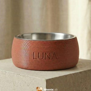 Può includere: Una ciotola per animali domestici color terracotta con interno in acciaio inossidabile. La parola "LUNA" è impressa sul lato. La ciotola è su un piedistallo quadrato di colore chiaro. Il logo WeenieLab è in basso a destra.