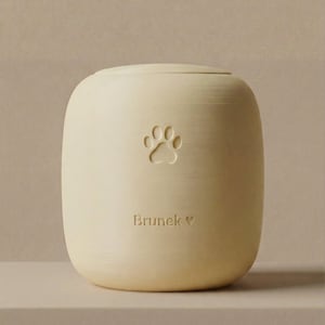 Puede incluir: Urna para mascotas de color crema con diseño de huella de pata. La urna tiene forma redondeada y tapa. El nombre "Brunek" está inscrito debajo de la huella, con un símbolo de corazón. La urna es de material sólido.