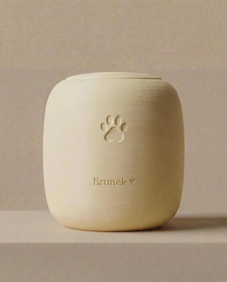 May include: A cream-colored pet urn with a paw print design. The urn has a rounded shape and a lid. The name "Brunek" is inscribed below the paw print, with a heart symbol. The urn is made of a solid material.