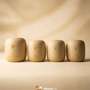 May include: Four beige pet urns with paw print designs. Each urn has a name and date engraved. The urns are arranged in a row against a neutral background. The WeenieLab logo is at the bottom.