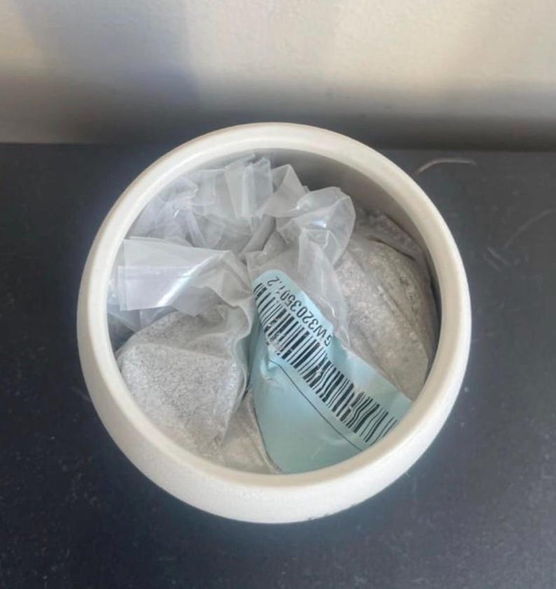 May include: A white, round ceramic container with a wide opening. Inside, there's a clear plastic bag filled with a gray, granular substance. A blue tag with a barcode and text is visible.