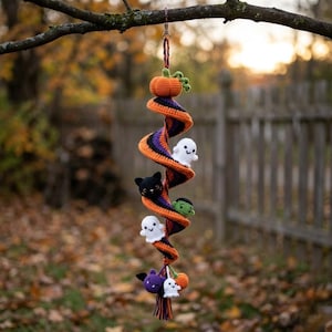May include: A spiral crochet hanging decoration featuring orange, purple, and black yarn. It includes small stuffed figures of a pumpkin, a black cat, a green monster, and white ghosts attached along the length, ending with a yarn tassel.