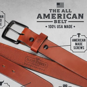 Op de afbeelding: Een close-up van een bruine leren riem met een zwarte gesp. De riem is gelabeld "The All American Belt" en "100% USA Made". De afbeelding benadrukt kenmerken zoals een Amerikaanse gesmede gesp, rundleer en in Amerika gemaakte schroeven en verpakking.