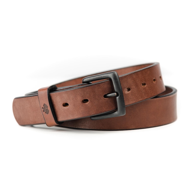 Op de afbeelding: Een bruine leren riem met een zwarte metalen gesp. De riem heeft een klassiek ontwerp met een rechthoekige gesp en verstelgaten. Het leer heeft een natuurlijke nerf en een subtiele glans. Een veelzijdig accessoire.