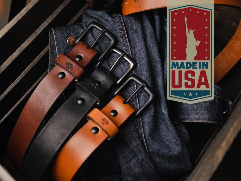 Op de afbeelding: Close-up van drie leren riemen in bruin- en zwarttinten met zwarte metalen gespen. De riemen zijn gerangschikt in een houten kist met een spijkerjasje. Een "Made in USA" label is zichtbaar.