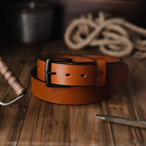 Op de afbeelding: Een opgerolde bruine leren riem met een zwarte gesp. De riem ligt op een houten oppervlak, met een verfroller en ander gereedschap op de achtergrond. De riem heeft een klassiek, eenvoudig ontwerp.