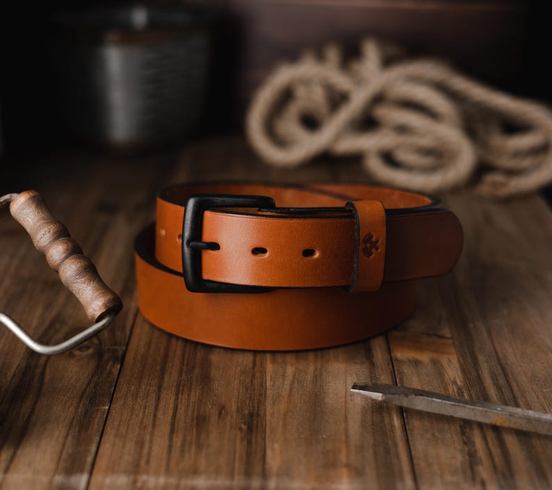 Op de afbeelding: Een opgerolde bruine leren riem met een zwarte gesp. De riem ligt op een houten oppervlak, met een verfroller en ander gereedschap op de achtergrond. De riem heeft een klassiek, eenvoudig ontwerp.