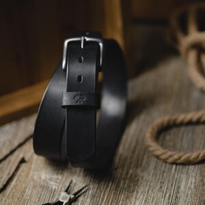 Op de afbeelding: Een zwarte leren riem met een zilveren gesp en een klein reli&euml;f embleem. De riem is opgerold op een houten oppervlak, met een rustieke uitstraling. De afbeelding bevat ook een touw en een metalen bak.