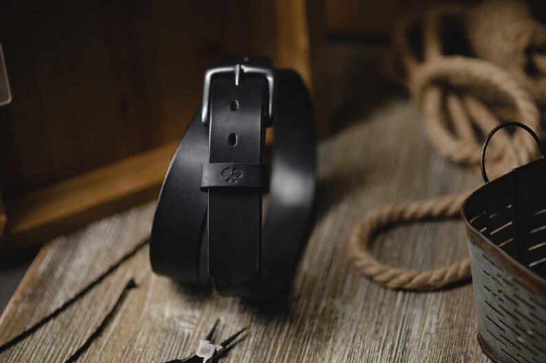 Op de afbeelding: Een zwarte leren riem met een zilveren gesp en een klein reli&euml;f embleem. De riem is opgerold op een houten oppervlak, met een rustieke uitstraling. De afbeelding bevat ook een touw en een metalen bak.