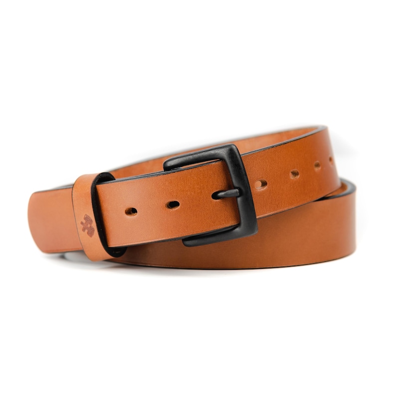 Op de afbeelding: Een bruine leren riem met een zwarte gesp. De riem is gemaakt van lichtbruin leer en heeft een zwarte metalen gesp. De riem is opgerold en heeft verschillende gaten voor aanpassing. De riem heeft een klein reli&euml;fontwerp bij de gesp.