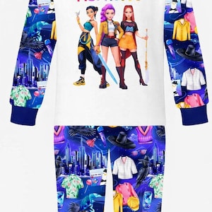 May include: White pajama set with long sleeves and pants. The top features the text "HUNTR/X" and a graphic of three women. The sleeves and pants have a repeating pattern of cityscapes, clothing, and other graphics. Blue trim.