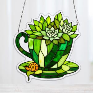 Könnte beinhalten: Glasmalerei mit einer Teetasse, die mit grünen Sukkulenten gefüllt ist. Die Tasse und Untertasse bestehen aus verschiedenen Grüntönen. Eine kleine Schnecke befindet sich auf der Untertasse. Das Kunstwerk wird von einer Kette gehalten.