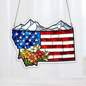 Puede incluir: Pieza de arte en vidrio manchado con la forma del estado de Montana, con el diseño de la bandera estadounidense. La bandera se combina con una cordillera y un arreglo floral. La pieza está diseñada para colgarse y se suspende mediante una cadena.