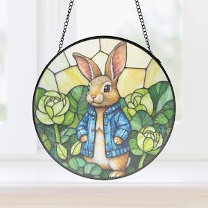 Può includere: Vetrata rotonda con un coniglio marrone con una giacca blu, circondato da vegetazione verde e gialla. L'opera d'arte è sospesa da una catena nera, ideale per una cameretta o come decorazione.