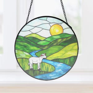 May include: Round stained glass panel featuring a pastoral scene with a white sheep, a blue river, and rolling green hills under a yellow sun. The artwork is framed in black and hangs from a chain.