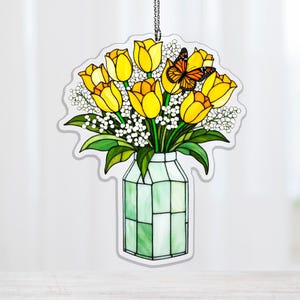 May include: A stained glass decoration featuring a bouquet of yellow tulips, white flowers, and green leaves in a green vase. An orange butterfly rests on the flowers. The decoration hangs from a silver chain.