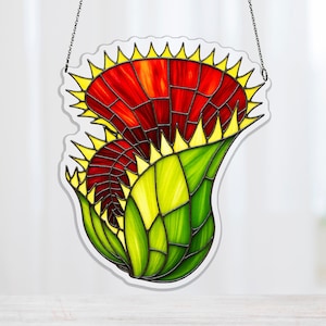 May include: Stained glass artwork of a Venus flytrap. The piece features a red and yellow flower head and a green and yellow base. The design is outlined in black and is suspended by a chain.