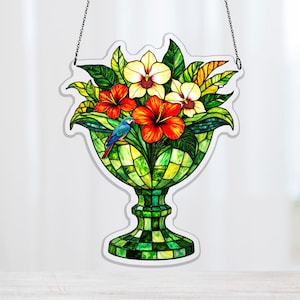 May include: A stained glass window hanging featuring a floral arrangement in a green vase. The arrangement includes red hibiscus, yellow orchids, and green leaves. A blue parrot is perched on the flowers. The vase has a checkered pattern.