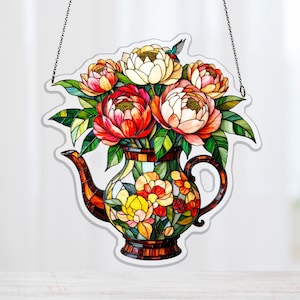 Puede incluir: Colgante decorativo de ventana de vidriera con forma de tetera. La tetera está llena de coloridas peonías y hojas verdes. Un pequeño colibrí está posado sobre las flores. El colgante de ventana está suspendido por una cadena.