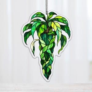 Puede incluir: Adorno decorativo de planta colgante con efecto vidriera. El diseño presenta una planta verde con hojas largas y fluidas y un tallo trenzado. Las hojas son de varios tonos de verde y amarillo, y el adorno cuelga de una cadena plateada.